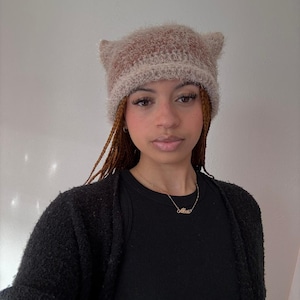 May include: A fuzzy, cat-ear beanie in shades of brown and beige. The hat is made of a soft, textured material. The beanie is a stylish accessory for cold weather.