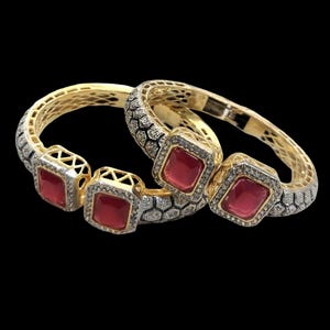 Peut inclure: Deux bracelets dorés avec des pierres rouges carrées et des diamants. Les bracelets ont un motif en nid d'abeille et sont posés sur un fond noir. Les pierres précieuses sont d'un rouge vif.