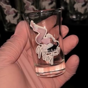 May include: A clear glass shot glass featuring a decal of a person in a light pink outfit with the text Nyaira's 21st written in a cursive script below the figure. The glass has a cylindrical shape with a thick base and smooth transparent walls.