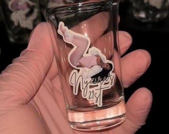 May include: A clear glass shot glass featuring a decal of a person in a light pink outfit with the text Nyaira's 21st written in a cursive script below the figure. The glass has a cylindrical shape with a thick base and smooth transparent walls.