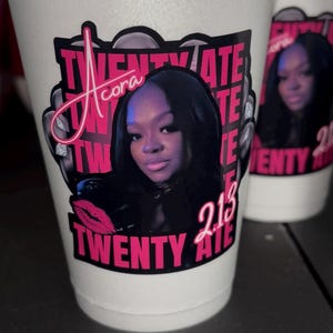 May include: A white foam cup featuring a custom graphic print with a portrait of a person, the name Acora in script, and the text TWENTY ATE 213 in bold pink lettering. The design includes a pink lip icon and a black background behind the portrait.