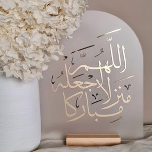 Op de afbeelding: Een boogvormig, mat acrylbord met gouden Arabische kalligrafie. Het bord wordt ondersteund door een rechthoekige lichtgekleurde houten voet. De kalligrafie luidt: اللهم اجعله منزلا مباركا. Het bord staat naast een witte vaas gevuld met witte hortensia's.