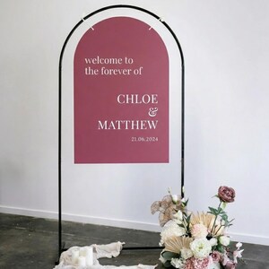 Op de afbeelding: Een welkomstbord voor een bruiloft met een bordeauxrood paneel met witte tekst: "welcome to the forever of CHLOE & MATTHEW 21.06.2024". Het bord staat in een zwart metalen frame, met bloemstukken aan de basis.