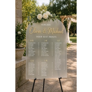 May include: An arched frosted acrylic wedding seating chart featuring the text 15.06.2025, Olivia & Michael, and YOUR SEAT AWAITS. The sign displays guest names organized by letter groups with table numbers, topped with white roses and green leaves.