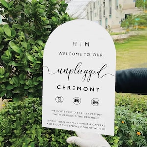 May include: A white sign with an arched top, displaying the words "HIM", "WELCOME TO OUR unplugged CEREMONY". The sign has icons for phones and cameras, and asks guests to switch them off.