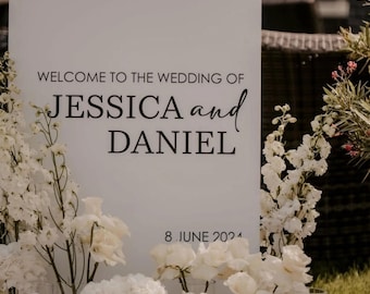 Puede incluir: Un letrero de boda blanco con texto negro que dice "WELCOME TO THE WEDDING OF JESSICA and DANIEL". La fecha "8 DE JUNIO DE 2024" está en la parte inferior. Decorado con flores blancas.
