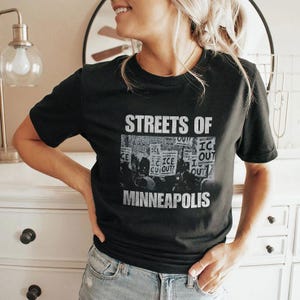 May include: Black t-shirt with the text "STREETS OF MINNEAPOLIS" in white. The shirt features a grayscale image of a protest with signs that read "ICE OUT!"