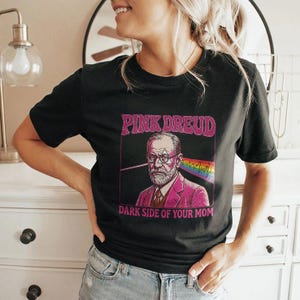 以下が含まれることがあります： メガネをかけた男性とピンクのスーツのグラフィックが特徴の黒いTシャツ。上部に「PINK DREUD」、下部に「DARK SIDE OF YOUR MOM」と書かれています。グラフィックには虹と線が含まれています。クルーネックです。