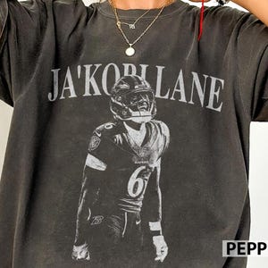 May include: A charcoal gray short-sleeve t-shirt featuring a distressed white graphic print of a football player wearing a helmet and jersey with the number 6. The text JA'KORIAN LANE is printed in a large arched font above the player graphic.