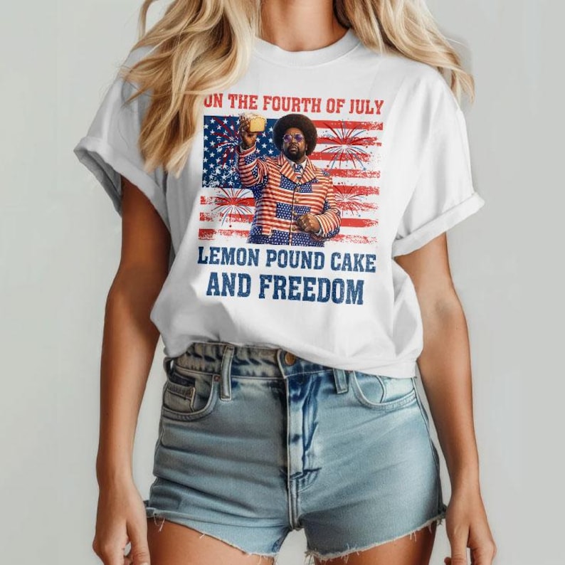May include: White t-shirt with a graphic featuring a man in a patriotic jacket, a flag, and fireworks. Text on the shirt reads "ON THE FOURTH OF JULY" and "LEMON POUND CAKE AND FREEDOM". The model is wearing denim shorts.
