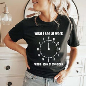 May include: Black crew neck t-shirt with white text that reads "What I see at work" above a clock graphic. The clock's hands point to the word "FUCK". Below the clock graphic, the text reads "When I look at the clock".