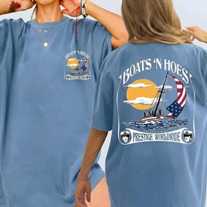 May include: A blue t-shirt featuring a graphic print of a sailboat with an American flag sail on the back and a smaller version on the front chest. The text reads BOATS 'N HOES and PRESENTED BY PRESTIGE WORLDWIDE with two small captain hat icons.