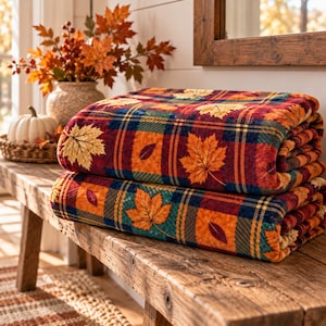 May include: A folded plush throw blanket featuring a vibrant autumn-themed pattern of red, orange, and yellow maple leaves layered over a dark blue, green, and red plaid background. The fabric has a soft, textured appearance with a repeating leaf motif.