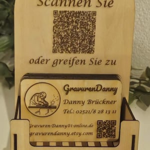 Könnte beinhalten: Ein hölzerner Visitenkartenhalter mit dem Text "Scannen Sie" und "oder greifen Sie zu". Der Halter zeigt das Logo "GravurenDanny" und Kontaktinformationen, einschließlich einer Website und Telefonnummer.
