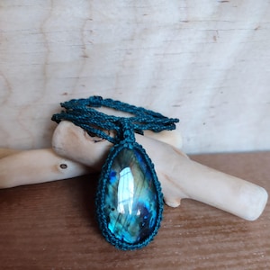 Op de afbeelding: Een ketting met een druppelvormige hanger van iriserende labradoriet. De steen is gezet in een turquoise macrame-omgeving en hangt aan een bijpassend koord. De ketting ligt op een houten ondergrond.