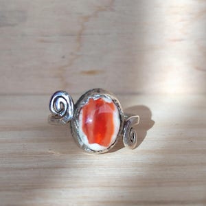 Könnte beinhalten: Silberner Ring mit einem ovalen, orange-weiß gefärbten Stein. Der Ring hat ein dekoratives Wirbeldesign auf beiden Seiten des Steins. Vor hellem Holzmaserungshintergrund.