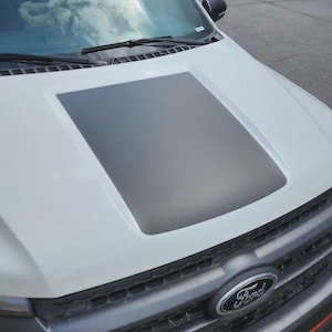 Op de afbeelding: Witte Ford-truckmotorkap met een donkergrijze rechthoekige accent. De motorkap heeft een zwarte ruitenwisser en een Ford-embleem. De afbeelding is genomen vanuit een iets verhoogde hoek en toont de voorkant van het voertuig.