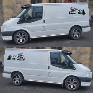 May include: Two white vans with black accents and custom wheels. Each van has a black graphic of a compass, mountains, and trees on the side. The vans are parked outside.