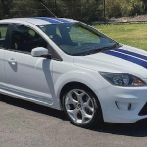 May include: White hatchback car with blue racing stripes on the bonnet and roof. The car has silver alloy wheels and is parked on a paved surface next to grass. The car has a spoiler.