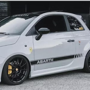 Op de afbeelding: Een lichtgrijze Abarth sportwagen met zwarte velgen en een zwarte streep met de tekst "ABARTH" op de zijkant. De auto heeft een zwart dak en gele remklauwen. Een klein geel embleem is zichtbaar.