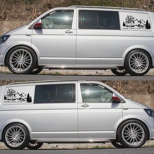 May include: Two silver vans with black decals showing a compass, mountains, and trees. The text "ONE LIFE LIVE IT" is below the graphic. The vans have black alloy wheels and are parked against a concrete wall.