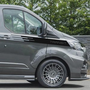 May include: A grey Ford Transit van with black side stripes and the word "TRANSIT" printed on the side. The van has a black front bumper and grey alloy wheels. The background features green foliage.