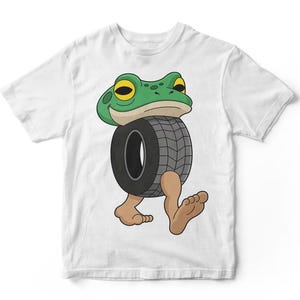 May include: White t-shirt featuring a cartoon frog with a green head and yellow eyes. The frog is depicted with a tire as its body and bare feet. The design is a playful and whimsical illustration.