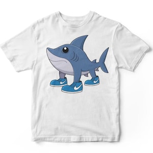 May include: White t-shirt featuring a cartoon shark with blue skin and blue sneakers. The shark has a friendly expression and is standing upright. The sneakers have white swoosh logos. The design is centered on the shirt.