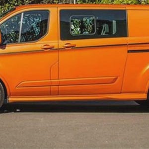 May include: Vibrant orange commercial van with black detailing and wheels. The van features a side door, tinted windows, and the word "Transit" is displayed on the side. The vehicle is parked on a paved road with green foliage in the background.