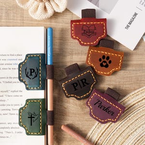 Puede incluir: Un conjunto de marcadores de cuero en varios colores, incluyendo burdeos, marrón y morado. Cada marcador presenta un diseño único, como una huella de pata, un libro o iniciales. También se ven un lápiz y un bolígrafo.