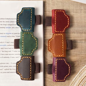 Puede incluir: Sujetalibros de cuero en varios colores: turquesa, verde, marrón, rojo, naranja y morado. Cada uno tiene un diseño cosido y una banda elástica. Los sujetalibros están sobre un libro.