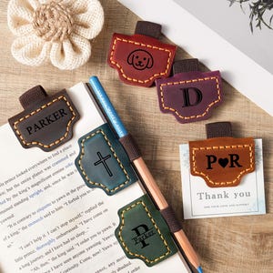Puede incluir: Marcadores de cuero en varios colores, incluyendo marrón, burdeos y verde, con bordes cosidos. Algunos presentan iniciales, un perro, una cruz y el nombre "Parker". También hay un lápiz y una tarjeta de "Gracias".