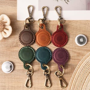 Puede incluir: Llaveros de cuero en varios colores: marrón, naranja, rojo, verde, turquesa y morado. Cada llavero tiene un diseño circular y un cierre metálico. Se muestran sobre una superficie de madera.