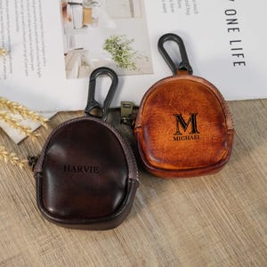 Peut inclure: Deux petites pochettes rondes en cuir avec fermetures éclair et mousquetons. L'une est marron foncé avec "HARVIE" en relief, l'autre est beige avec "M MICHAEL" en relief. Les deux ont un aspect vintage et usé.