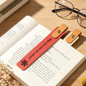 Puede incluir: Un marcapáginas de cuero rojo con el texto impreso en negro 'May success be yours!' y un icono de copo de nieve, rematado con una lengüeta de cuero beige y un botón de presión metálico color latón, sobre la página de un libro abierto.