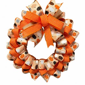 May include: A circular wreath constructed from tan fabric ribbon featuring a repeating pattern of black, white, and orange pumpkins. The wreath is accented with a large, bright orange bow with white polka dots and a burlap-textured center ribbon.