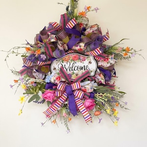 Pode incluir: Uma grinalda decorativa com um design floral e de fita. A grinalda apresenta um letreiro que diz "Welcome" e é rodeada por fitas rosa, roxas e brancas, e flores coloridas. A grinalda foi concebida para ser pendurada numa porta ou parede.