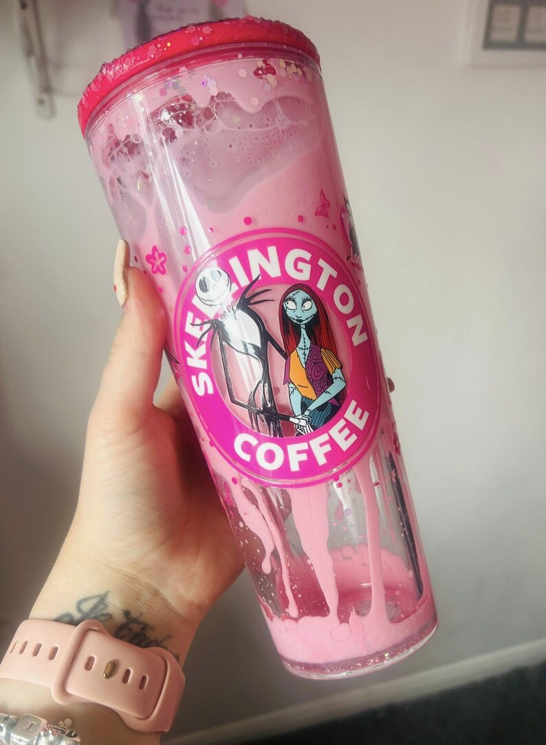 May include: A pink tumbler with a screw-on lid, filled with a pink liquid. The tumbler features a circular graphic with Jack Skellington and Sally from The Nightmare Before Christmas, with the text "SKELLINGTON COFFEE".