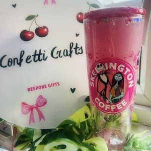 May include: A pink tumbler with a glittery rim and a graphic of Jack Skellington and Sally from The Nightmare Before Christmas. The tumbler has a "Skellington Coffee" logo. A white decorative plate with cherries and the text "Confetti Crafts" is in the background.