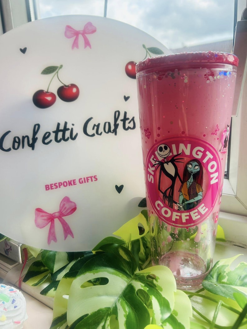 May include: A pink tumbler with a glittery rim and a graphic of Jack Skellington and Sally from The Nightmare Before Christmas. The tumbler has a "Skellington Coffee" logo. A white decorative plate with cherries and the text "Confetti Crafts" is in the background.