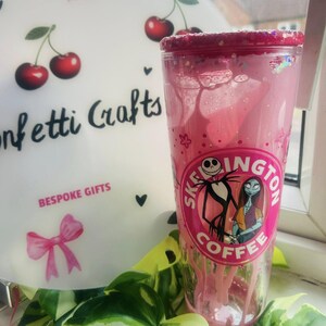 May include: A pink tumbler with a glittered rim featuring a design of Jack Skellington and Sally from The Nightmare Before Christmas. The tumbler has the text "Skeington Coffee" and is surrounded by green leaves.