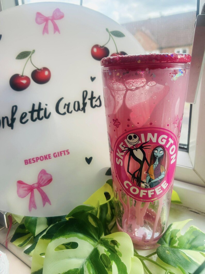 May include: A pink tumbler with a glittered rim featuring a design of Jack Skellington and Sally from The Nightmare Before Christmas. The tumbler has the text "Skeington Coffee" and is surrounded by green leaves.