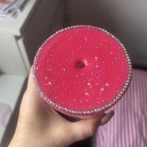 May include: A close-up of a pink tumbler with a sparkly, glittery finish. The lid is adorned with a ring of rhinestones. The tumbler has a hole for a straw. The background is blurred.