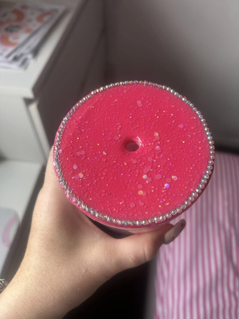 May include: A close-up of a pink tumbler with a sparkly, glittery finish. The lid is adorned with a ring of rhinestones. The tumbler has a hole for a straw. The background is blurred.