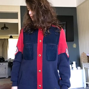 May include: A navy blue and red jacket with gold buttons. The jacket features two front pockets and a red central panel. The sleeves have a white and red emblem. The bottom of the jacket has an embroidered design with the text "ERIC".