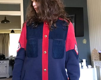 Pode incluir: Casaco azul marinho e vermelho com botões dourados. O casaco tem dois bolsos frontais e um painel central vermelho. As mangas têm um emblema branco e vermelho. A parte inferior do casaco tem um desenho bordado com o texto "ERIC".