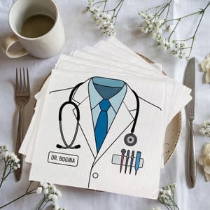 May include: White paper napkins with a doctor's coat design, including a stethoscope, blue tie, and the name "DR. BOGINA." The napkins are stacked on a plate, with a fork, knife, and a cup in the background. Decorative flowers surround the plate.