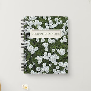 Op de afbeelding: Een spiraalgebonden notitieboek met een omslag voorzien van een dicht patroon van kleine witte bloemen en groen blad. Een rechthoekig label in het midden bevat de tekst "JOURNALING MY LOSS" in een schreefloos lettertype op een crèmekleurige achtergrond.