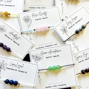 Può includere: Assortimento di braccialetti con pietre preziose con carte dei segni zodiacali. Ogni braccialetto presenta perline di diversi colori, tra cui rosa, viola, verde e blu. Le carte mostrano il nome della pietra preziosa, le date di nascita e i simboli zodiacali. Sfondo bianco.