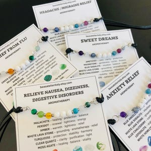 Op de afbeelding: Assortiment van aromatherapiearmbanden met verschillende edelsteen kralen, elk bevestigd aan een zwart koord. Elke armband wordt geleverd met een kaart met het doel, zoals hoofdpijnverlichting of angstverlichting, en de gebruikte edelstenen.